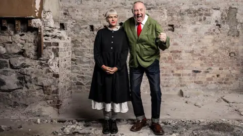 Patch Dolan Keith and Marj stood next to one another. Marj is looking a Keith out the corner of her eye. Keith has his fist clenched and mouth open with an excited expression.