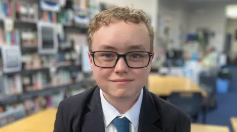 Rory has curly blonde hair, is wearing black rimmed glasses and is wearing a blue blazer, white shirt and blue and yellow tie. She is sitting in a classroom full of books.
