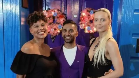Bored of Dating Apps Two women and a man smiling at the camera. They are standing in front of a blue wall with two pink wreaths on. The women are both wearing black tops. The man is wearing a purple jacket and matching waistcoat over a white T-shirt.