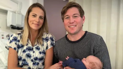 Emma Baugh/BBC Jessica Williams with below shoulder-length fair hair, wearing a white top with large blue flowers. Sitting beside her is her husband Pete, with short light brown hair, wearing a black and white jumper. Both are smiling towards the camera. He is holding their son Orlando, who is sleeping and wrapped in a dark blue knitted cardigan.