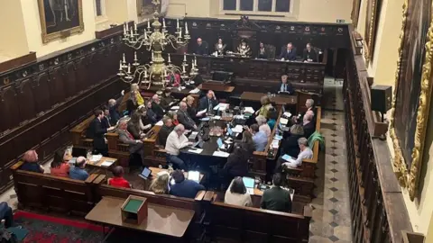 Exeter City Council members sitting in the Guildhall in Exeter with the Mayor of Exeter seated in front of a mace overseeing the meeting.