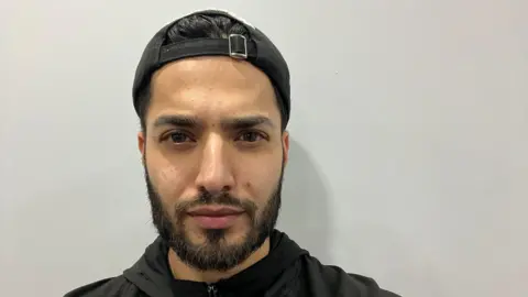 Sakib Hussain Sakib Hussain, a man with a short black beard is standing against a light-coloured wall. He is wearing a black baseball cap backwards and a black top.