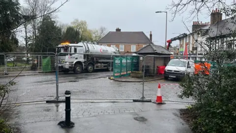 Tony Fisher/BBC A tanker clearing water in Chalfont St Peter. It shows a road cordoned off by a metal fence, a tanker in the distance and a cone.