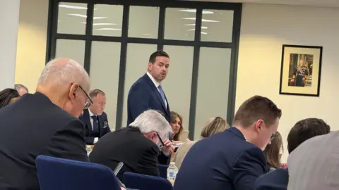 Simon Dedman/BBC Gavin Callaghan is in the centre of the frame, standing and talking, at a council meeting. He is wearing a navy blue suit and blue tie. Other people - mostly men - are sitting down and wearing suits.