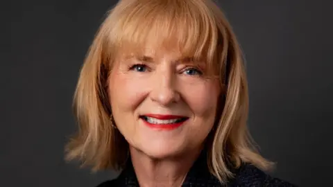 Caroline Biggs Caroline Biggs, who has shoulder-length blonde hair and a fringe to her eyebrows. She is wearing a just-seen black jacket and is smiling. 