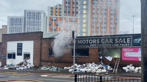 Smoke is billowing out of a building. Next to it, or part of the same building, is a black sign which says 'KM Motors Car Sale and Used Cars You can trust.' 
