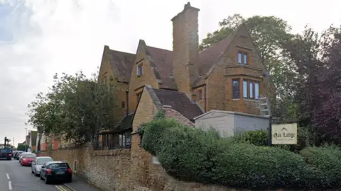 Google Brick-built three-storey care home surrounded by hedges on a road with cars parked.
