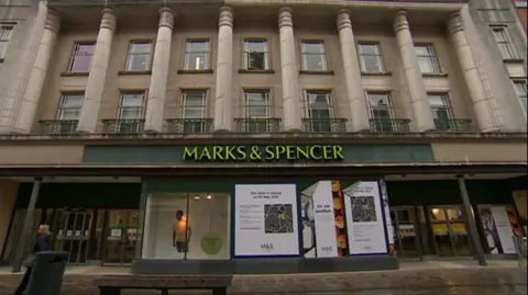 The exterior of the former Marks and Spencer store in Hull. A green sign is placed above the doors of the building, which has three floors and ornate stone pillars on the facade. The windows on the ground floor are covered by large blue and white graphics with information about the store closure.