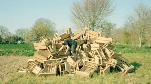 Alexander Mourant, a young man in plain dark clothes, climbing in what appears to be a large pile of wooden pallets in a green field.