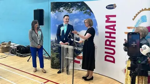 Darren Grimes, centre, on election night wearing a suit with a light blue tie and light blue and white Reform UK rosette. His is tall with dark hair. Karen Allison stands next to him. She had long dark hair and wears blue trousers. Journalists are next to them, as well as a woman who is announcing the election results