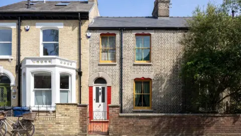 Matthew Smith The exterior of a terraced house made of yellow and brown bricks, with yellow painted windows trimmed in red. It is attached on either side, with the house on the right covered by a large tree in leaf. The house on the left is also built of yellow brick and has a white-painted bow window. 