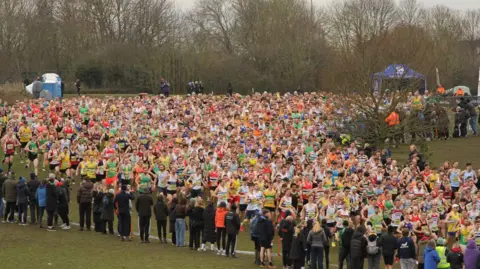 A crowd of runners in different coloured running vests running down a hill. There are spectators lining either side of them. They are on a grassy bank which is lined with leafless trees in the background.