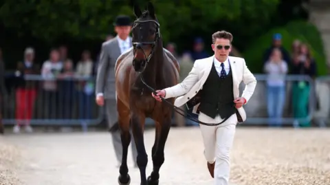 A smart young man, in a white suit with a black waistcoat and blue tie, running along a path whilst holding the reins of a a race horse. There is a man in a grey suit and bowler hat behind him and railings with spectators standing in the background