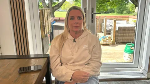 Lisa Barker sits in a living room in front of a patio door. She has shoulder-length blonde hair and is wearing a cream hoodie. Her mobile phone is on the table next to her. 