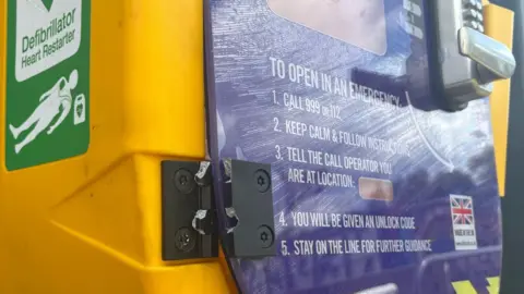 The black hinge of the yellow and purple wall-mounted defibrillator box has been snapped. The front of the box has instructions on how to use the equipment and a code-protected metal door handle.