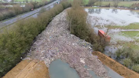 A large pile of waste next to a field and road. The waste stretches into the distance between two rows of trees.