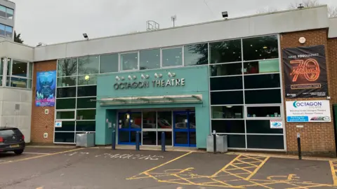 The outside of the theatre, which is a low two-storey building. It has panes of glass windows around entrance, which is turquoise and has the words 'octagon theatre' above the door. 