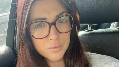 Charlotte Angel, who has long auburn hair and wears large red-framed glasses, poses for a selfie in the front seat of her stationary car.