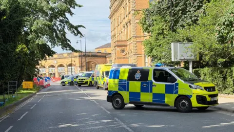 Three police vans and an ambulance parked on a closed road with traffic cones up one side. In the distance a station building.
