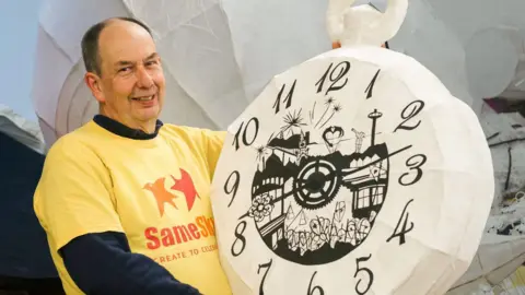 A man in a yellow t-shirt holding a papier-mache clock.