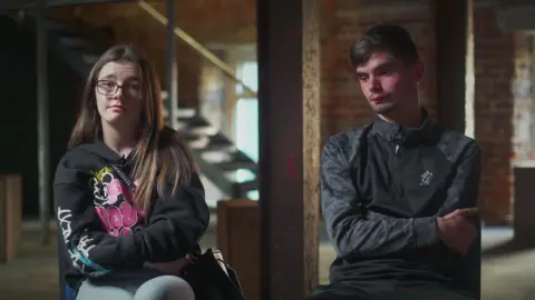 Kenzie and John sit on chairs in a large empty warehouse. She has brown hair, round glasses and is wearing a black jumper with bright coloured writing. John has dark hair and is wearing a black quarter zip jacket with grey sleeves.