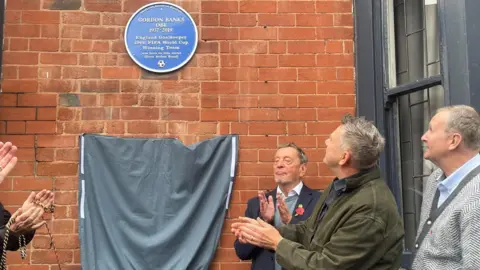 Simon Thake Three men look up and applaud a blue plaque displayed on a brick wall. A grey curtain drops below it.