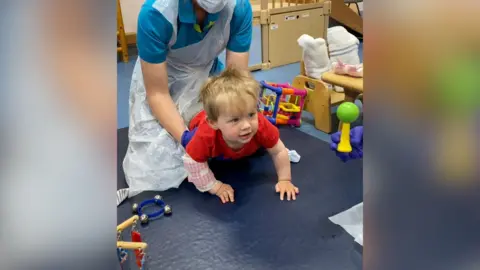 A child wearing a red t-shirt is crawling on the floor with a piece of fabric wrapped around his right arm. A nurse is behind him helping to hold him up.