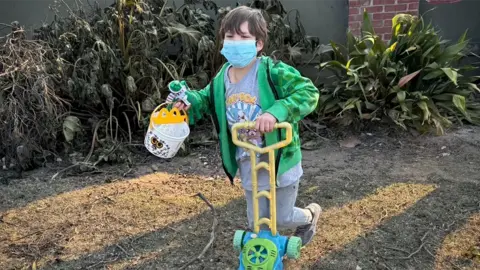 A boy holds a bucket in his right hand and carries a brightly coloured toy lawnmower in his left