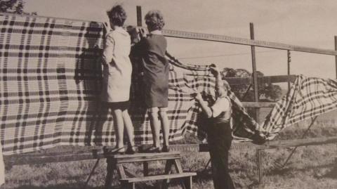 Black and white picture of two women putting up patterned wool cloths on a frame in the open air