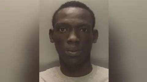 West Midlands Police A mugshot of a man with short black hair and wearing a grey jumper, standing against a wall.