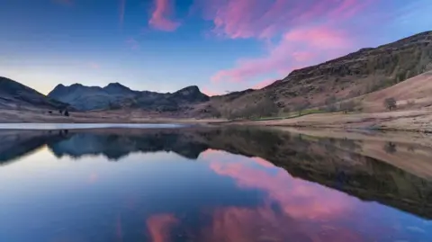 Jonny Gios View of the head of a lake fringed by mountains, which are reflected in the water. The sky is blue with clouds denoting sunset or sunrise, and these are also reflected.