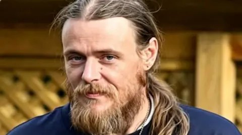Sean Forster, a man in his late 30s, with shoulder-length, blonde hair and a long beard. He is smiling slightly.