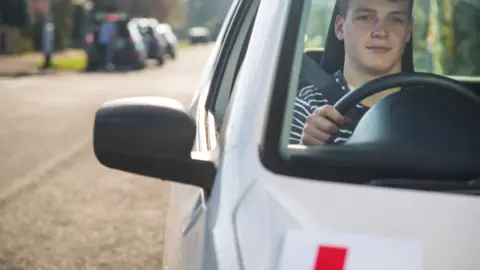 Young man driving a silver car with a red Learner badge on the front