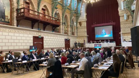 West Northamptonshire Councillors sat at desks with microphones with several people sat up on a stage