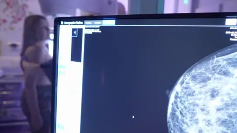 An X-ray image of a breast on a screen and a seated woman is blurred in the background