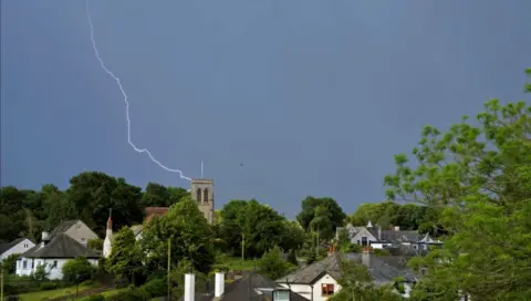 JL Park Photography Thunder storm coming in over the bay into Silverdale, Lancashire. Lightning can be seen above a church tower, with peaked roof houses coming down the hill
