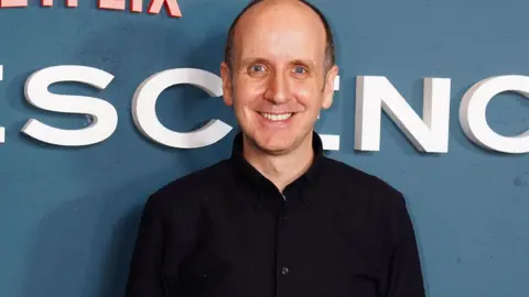 PA Media Jack Thorne standing in front of a blue wall at the premiere screening of Adolescence. He is wearing a black shirt and is smiling at the camera.