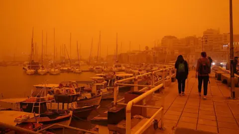 Stefanos Rapanis/Anadolu via Getty Images People walk along a waterfront with boats, buildings and the sea in the background. The sky is orange.
