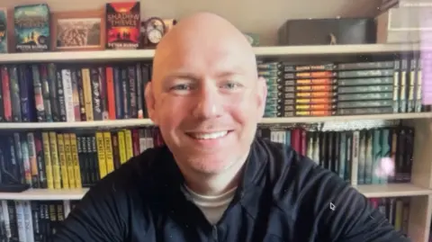 Peter Burns sits at home in his study with copies of his book Shadow Thieves on the bookshelf in the background. He is bald and wears a black shirt with white t-shirt underneath. 