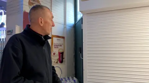 A man standing next to white metal shutter doors, which have been brought down over a shop doorway. He is wearing a black zipped fleece. On the wall behind him, a poster advertises takeaway coffee.