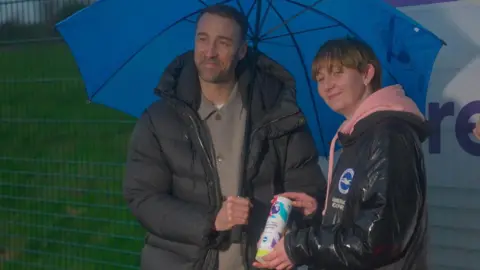 Glenn Murray hands the award which is multicoloured and barrel shaped. Both are wearing raincoats and Glenn is holding an umbrella