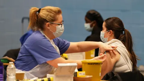 PA Media A healthcare worker in blue scrubs, a face mask and glasses administers a vaccine into the upper arm of a young woman wearing a white top. They are seated at a table with medical supplies including a yellow sharps bin. Another healthcare worker in a face mask can be seen in the background against a light blue wall.

