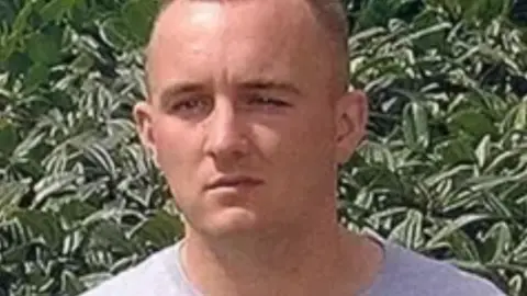Daniel Keatinge, a clean-shaven man in his mid-20s with short light blonde hair, wearing a light coloured t-shirt, looks into the camera in front of a large green hedge or bush. 