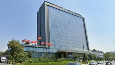 A large, modern, rectangular, grey building with Turkish flags outside