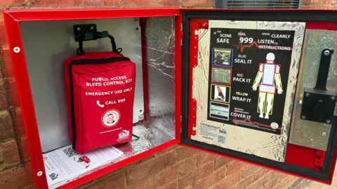 An opened red emergency bleed control box mounted on a brick wall, showing a red zip-up kit inside labelled “Public Access Bleed Control Kit – Emergency Use Only – Call 999”. The box interior is lined with reflective silver insulation material. On the inside of the door is a large instruction panel with diagrams outlining steps for treating severe bleeding, including calling 999 and using items. A keypad entry system is visible on the right side of the box for controlled access.