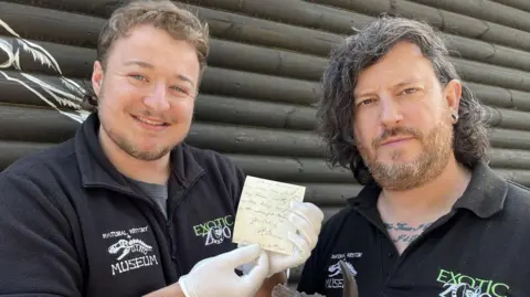 Exotic Zoo Two men pictured in black polo shirts with Exotic Zoo written on the breast. The man on the left has light brown curly short hair and a light beard and moustache. He smiles as he holds a small yellow fragment of paper the size of a post-it note. He is wearing white gloves. The man on the right has a tattoo just visible across the base of his neck, long black curly hair in a side parting, a greying goatee and an earring in his left ear. The pair stand outside a black wooden hut.
