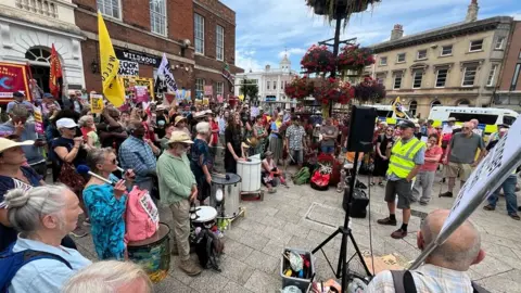 Counter-demonstrators gathered around a set of speakers in a town square. Some of them have drums and are holding placards.