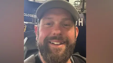 James Desborough is smiling. He has a brown beard and is standing in front of a blue tractor. He has a grey baseball cap on.