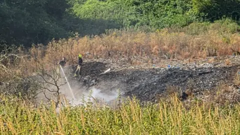 In the centre of the image, which was taken from far away, two firefighters in uniforms and fluorescent yellow helmets are visible. They are standing on top of a tall mound of rubbish, the size of which is akin to what one would expect at an official landfill site. One firefighter is using a large handheld hose and water is coming out of it towards the ground in front of them which is smouldering and emitting smoke. In the foreground and background are thick shrubs, hedges and trees signalling it is a rural location.
