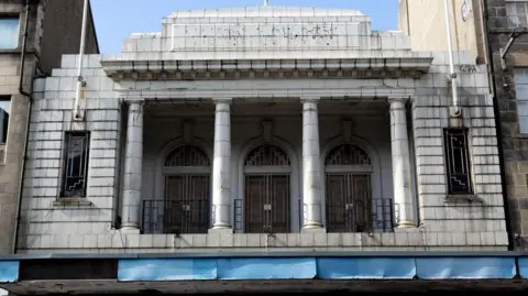 Anja Wolf The former cinema is built of white blocks, with four stone pillars at its entrance.